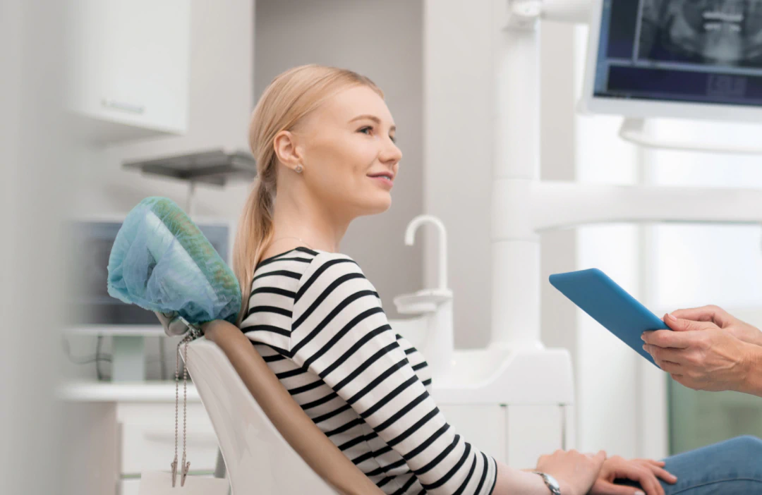 A woman sits in a dental chair, smiling and looking at a person holding a blue tablet. Dental equipment and a computer screen are visible in the background.