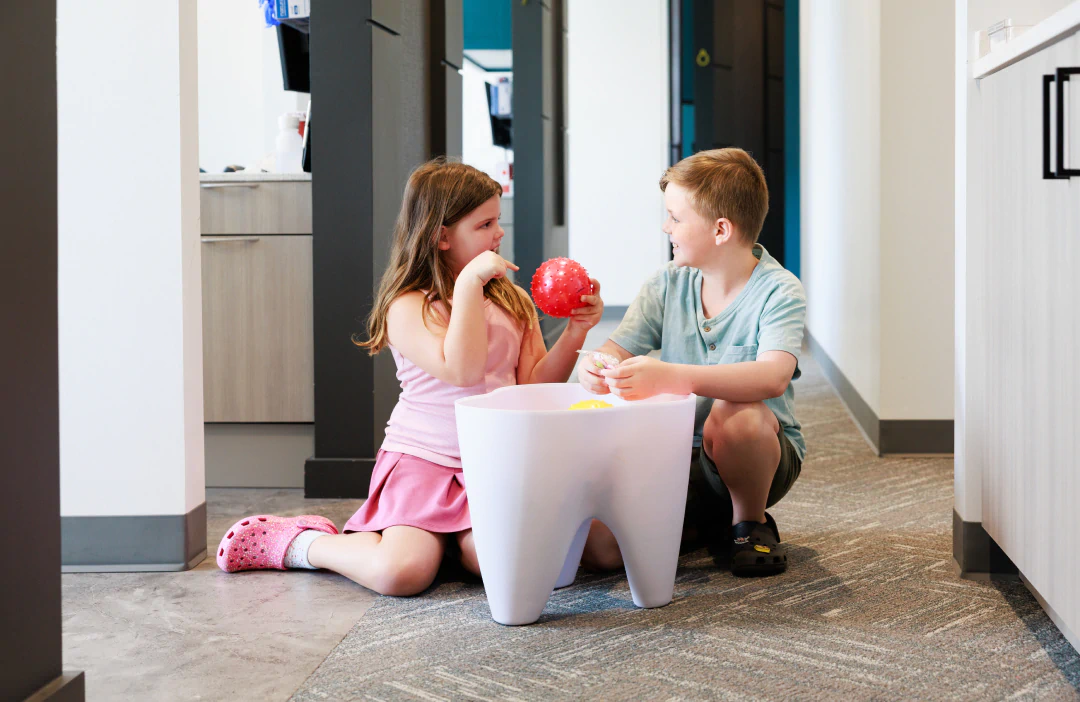 Two children sit on the floor in a hallway, playing with toys next to a large container shaped like a tooth. The girl holds a red ball, and both are smiling and interacting with each other.