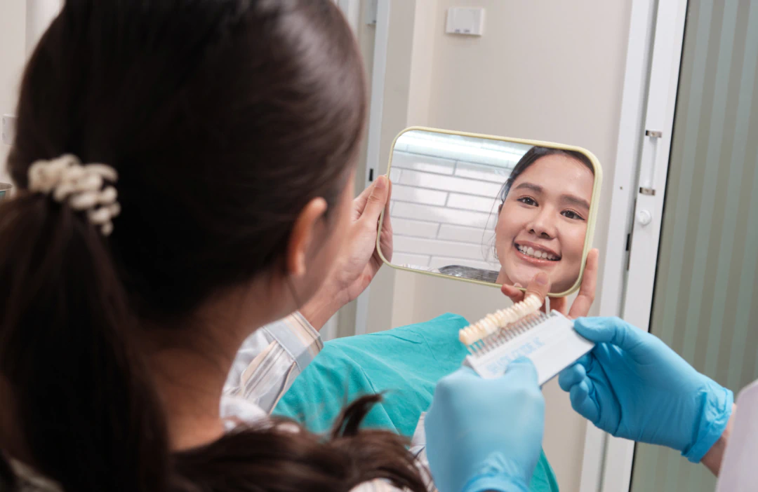 A woman sitting in a dental chair smiles at her reflection in a handheld mirror while a gloved dentist holds a tooth shade guide near her mouth.