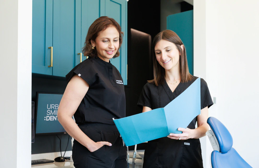 Two women in black medical scrubs smile while reviewing a blue folder in a modern dental office. Blue cabinets and a dental chair are visible in the background.