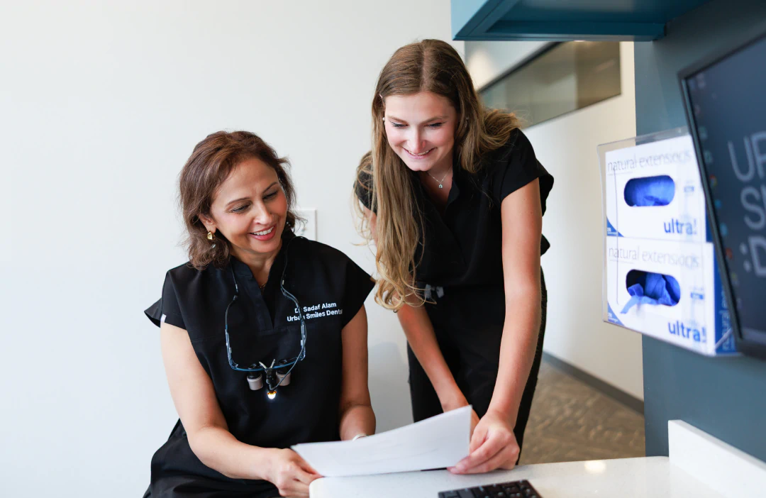 Two women in black scrubs smile and look at a document together in a dental office. One is seated with dental loupes, while the other stands, leaning over the desk. A box of gloves is visible on the wall.