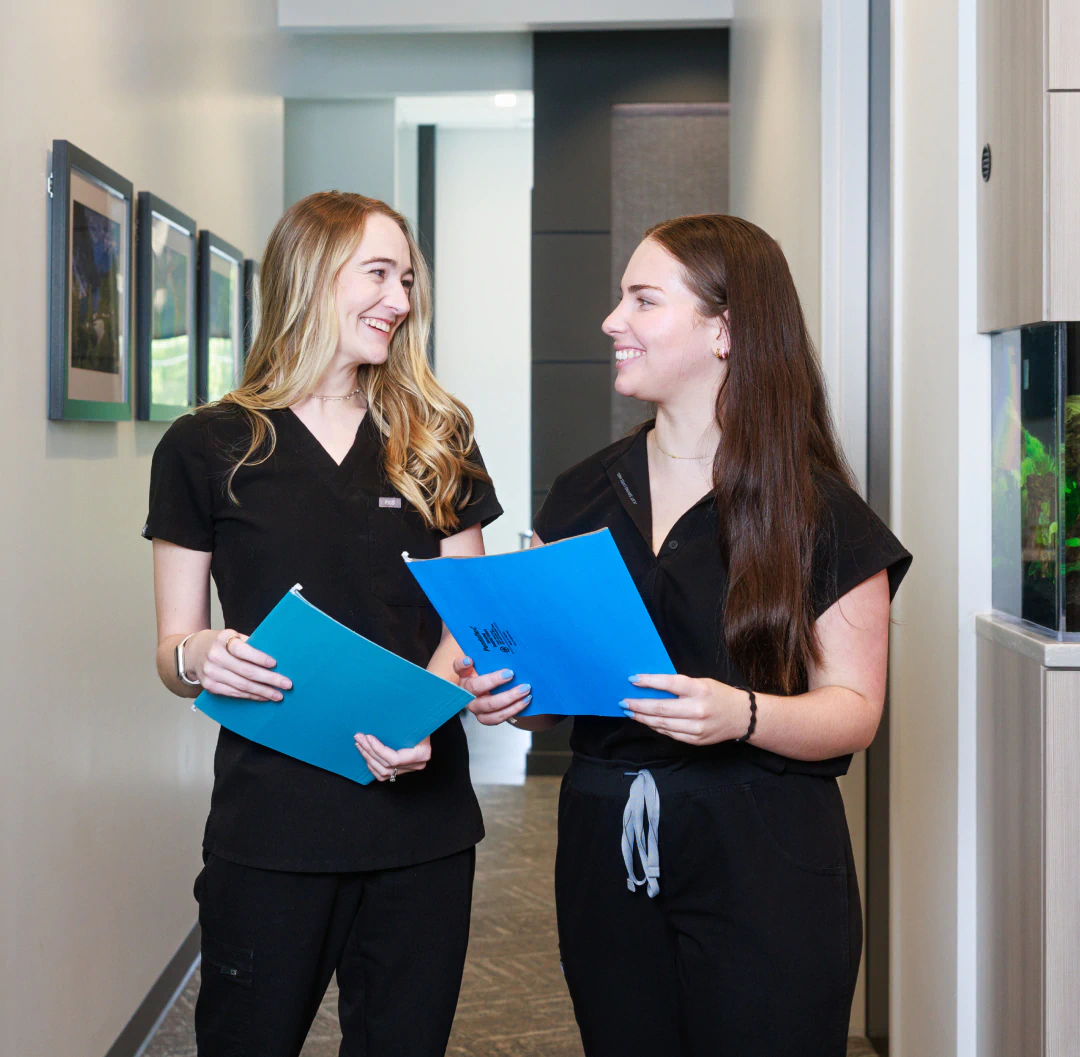Two women in black scrubs stand in a hallway, holding blue folders and smiling at each other. Art hangs on the wall behind them, and theres a fish tank to the right.