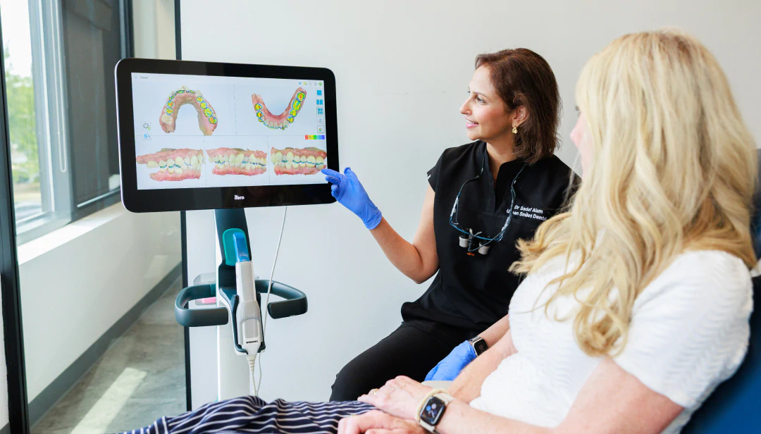 A dental professional shows a patient digital images of teeth on a screen, explaining details while both are seated in a modern clinic.