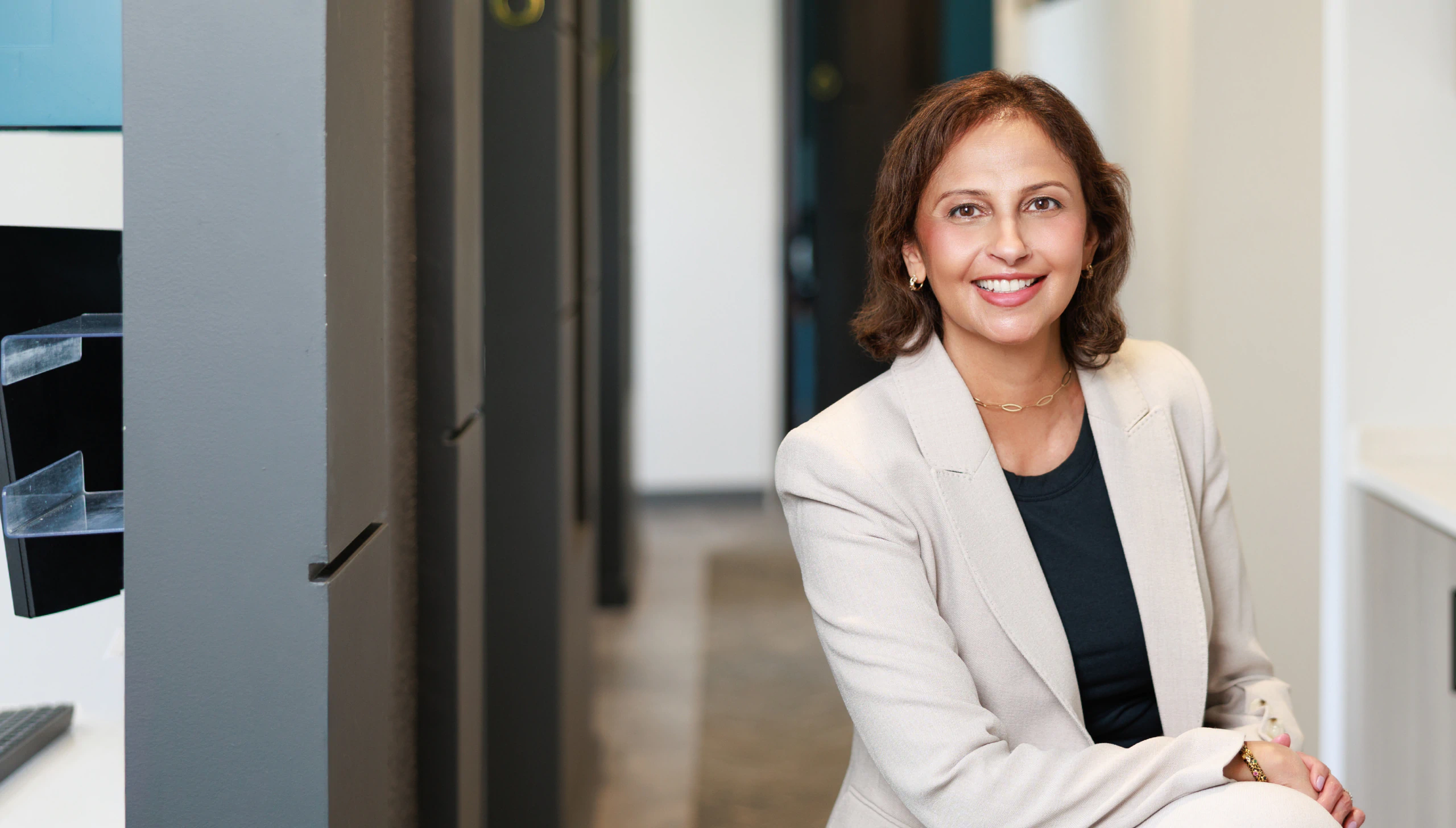Mobile_Home_Your_Partner_In_Health_Background A woman in a light blazer and black top smiles while sitting in a modern office hallway with gray walls and a blurred background.