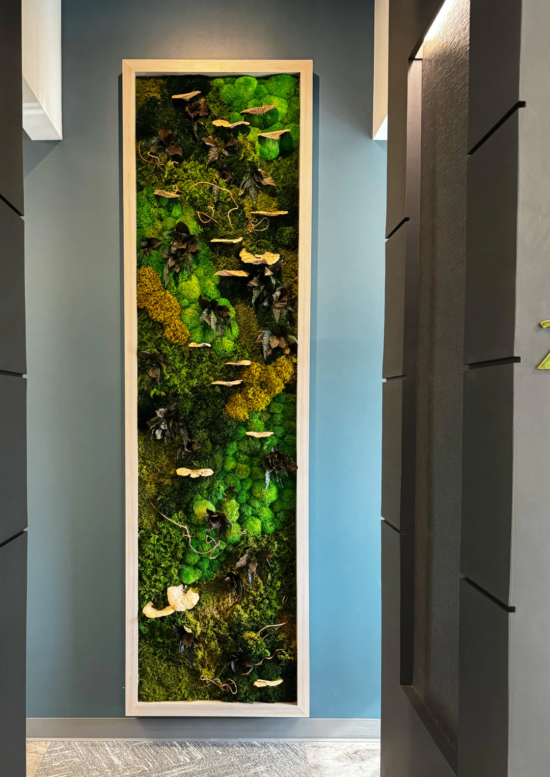 A tall, rectangular wall art piece features vibrant green moss, yellow moss, and dark leaves arranged vertically in a wooden frame, mounted on a blue wall.