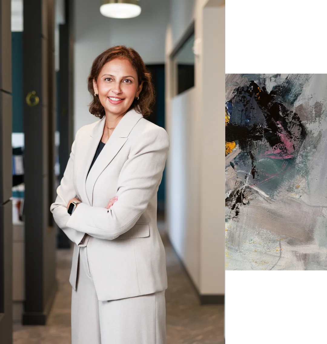 A woman in a light-colored suit stands smiling with arms crossed in an office hallway; abstract art is visible on the right side of the image.