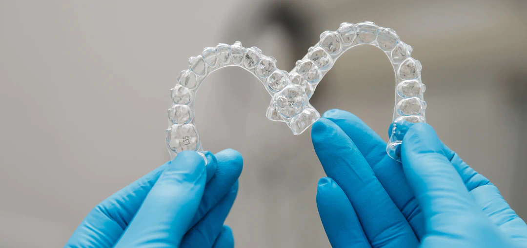 Two gloved hands hold clear dental aligners shaped like the letter M against a blurred background.