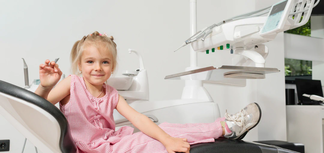 A young girl in a pink striped outfit sits in a dental chair, smiling and waving, inside a modern dentists office with dental equipment in the background.