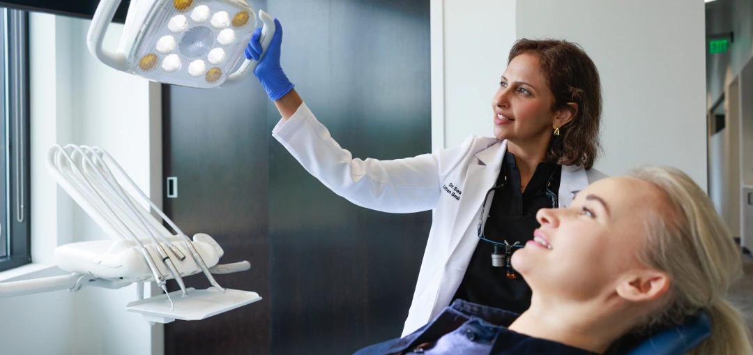 A dentist adjusts an overhead light while a patient reclines in a dental chair, preparing for an exam in a modern dental office.