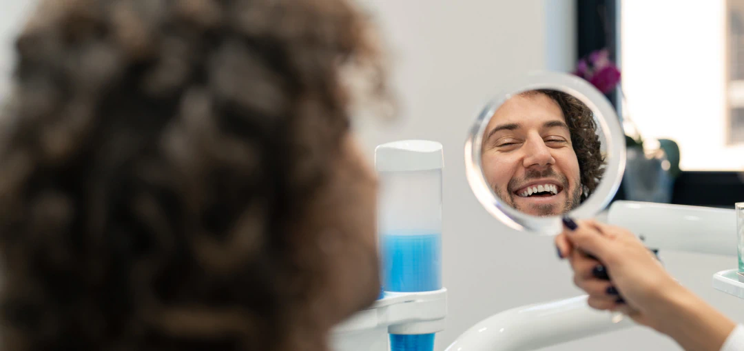 A person with curly hair smiles while looking at their reflection in a handheld mirror at what appears to be a dental clinic.